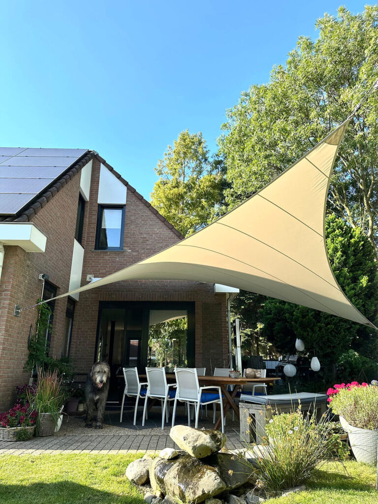 Outdoor dining area in a brick house yard with a large beige tensile sail shade and white chairs, a dog nearby.