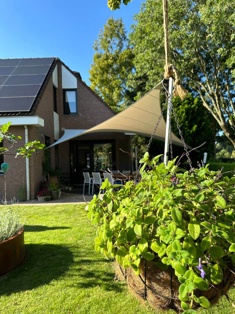 House with solar panels on the roof, a white shade sail over a patio with chairs, and a hanging plant basket in the foreground on a sunny day.