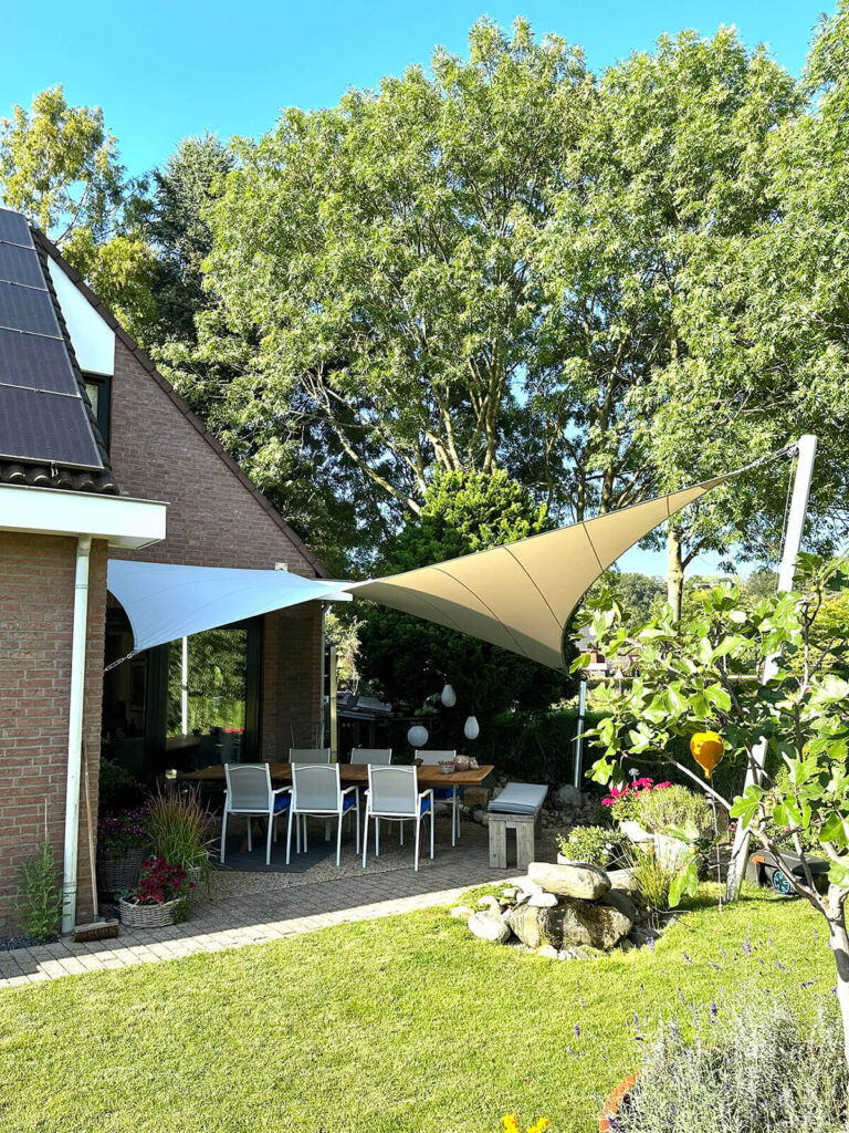Outdoor dining area in a sunny backyard: a wooden table with white chairs under white and beige shade sails, against a brick house with a lush green lawn and large trees beyond.