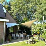 Rear patio scene: brick house with solar panels, outdoor table and white chairs under sail shade, surrounded by trees and garden.