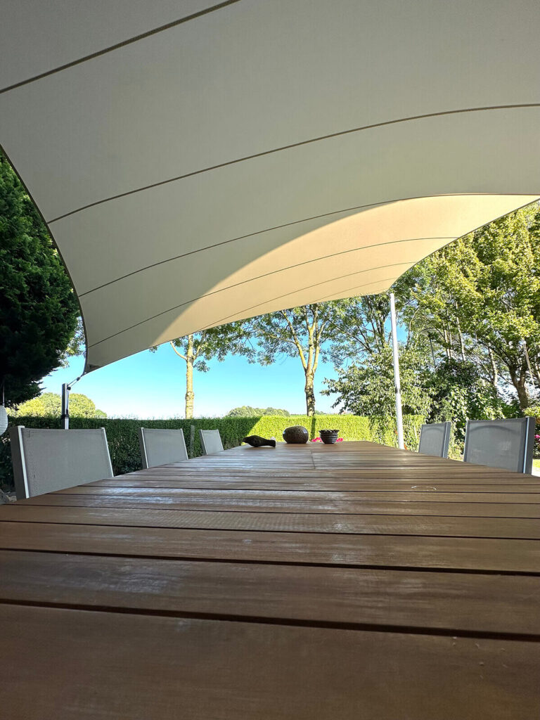 Long wooden outdoor table under a curved beige shade sail with white chairs and green hedges behind.