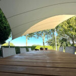Outdoor dining table under a curved shade sail with wooden slats and white chairs, backyard greenery in the background