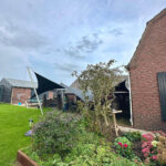 Backyard with brick house, two dark shade sails over a patio, a person near outdoor seating, and a flowerbed in the foreground.