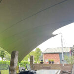 Outdoor patio scene shaded by a large curved fabric canopy, with a glass-top table and metal chairs beneath it.