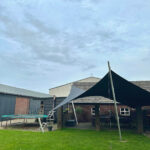 Backyard scene with a trampoline on the left, metal ladder, and a patio area under dark and light-gray shade sails tied to posts next to a brick house under a cloudy sky.