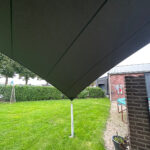 Large dark shade sail stretched over a backyard, supported by a metal pole, with a lawn, hedges, and a brick building nearby.