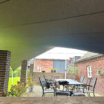 Outdoor patio under a slanted roof with a dining table and chairs, brick pillars, and string lights in a backyard setting.