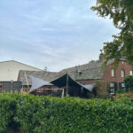 Backyard patio with triangular navy and light gray shade sails, green hedges, and a brick house under a blue sky.
