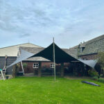 Backyard patio with a large black triangular shade sail anchored to a central pole, covering seating area beside a brick house.