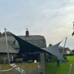 Backyard scene with two large shade sails (black and gray) stretched between metal poles in front of a brick house; a person stands on the lawn to the right.