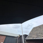 Backyard scene under a large black shade sail with a trampoline in the foreground and brick buildings in the background.
