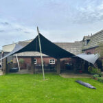 Backyard with a large dark triangular shade sail supported by metal poles, brick house behind, and a green lawn front.
