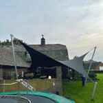 Backyard scene with a large trampoline in the foreground and black and gray shade sails stretched between metal poles over it.