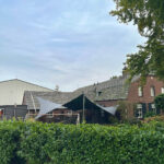 Backyard scene with a brick house, green hedge in the foreground, and black and white triangular shade sails over a patio area under a blue sky.