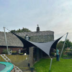 Backyard with a large black and gray shade sail attached to tall metal poles in front of a brick house.