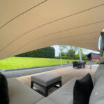 Outdoor patio with a large beige curved canopy over a cushioned seating area and a distant dining table on tiled ground against a lawn and trees. A small dog sits near the table.