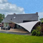 Backyard scene with a brick house and tiled roof, a large white triangular shade sail over an outdoor dining area on a tiled patio.