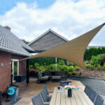 Outdoor patio with a large beige shade sail over a dining table and lounge seating, brick house and garden in the background.