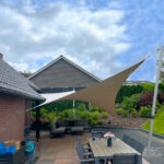 Backyard patio with a large beige shade sail over a dining table and chairs, brick house and garden in the background.