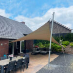 Backyard patio with a beige triangular shade sail over a dining table and grey outdoor seating next to a brick house.
