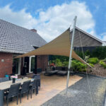 Backyard patio with beige shade sail anchored to a metal pole over outdoor dining table and lounge seating.