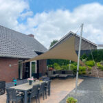Backyard patio with a long grey dining table and chairs beneath a beige shade sail, brick houses in the background.
