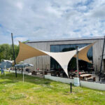 Outdoor cafe with beige and white tensioned sun sails in front of a wooden modern building, picnic tables, and umbrellas under a partly cloudy sky.