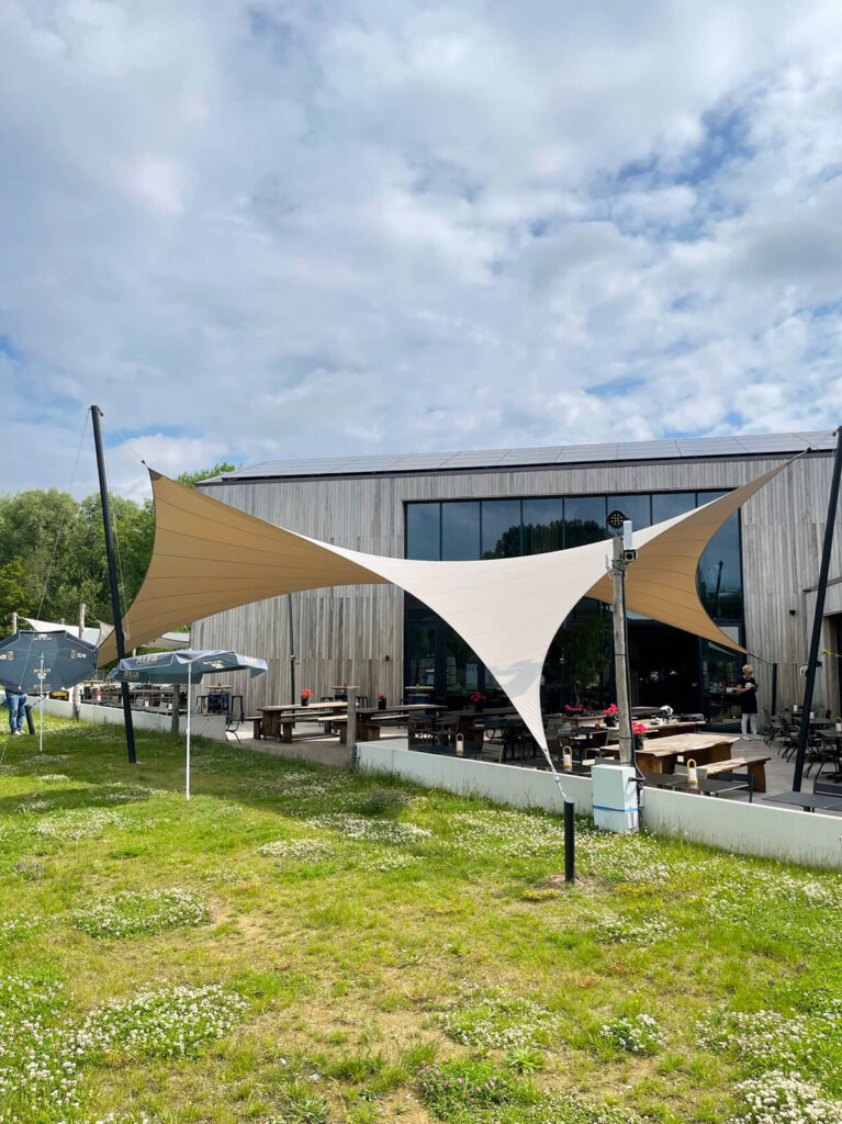 Outdoor cafe with beige and white tensioned sun sails in front of a wooden modern building, picnic tables, and umbrellas under a partly cloudy sky.