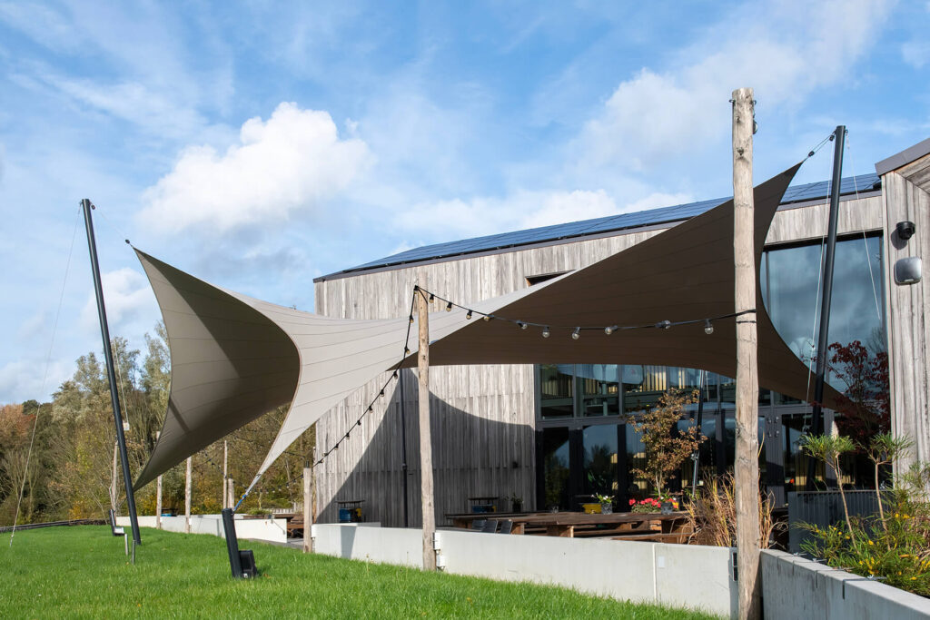 Modern wooden-clad building with large beige fabric shade sails and string lights over a green lawn.