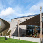 Modern wooden-clad building with large beige fabric shade sails and string lights over a green lawn.