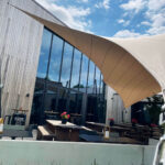 Outdoor cafe patio with wooden picnic tables under a curved beige sunshade beside a glass-fronted modern building.