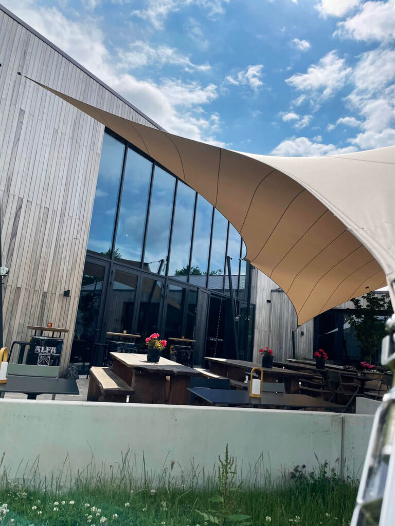 Outdoor cafe patio with wooden picnic tables under a curved beige sunshade beside a glass-fronted modern building.