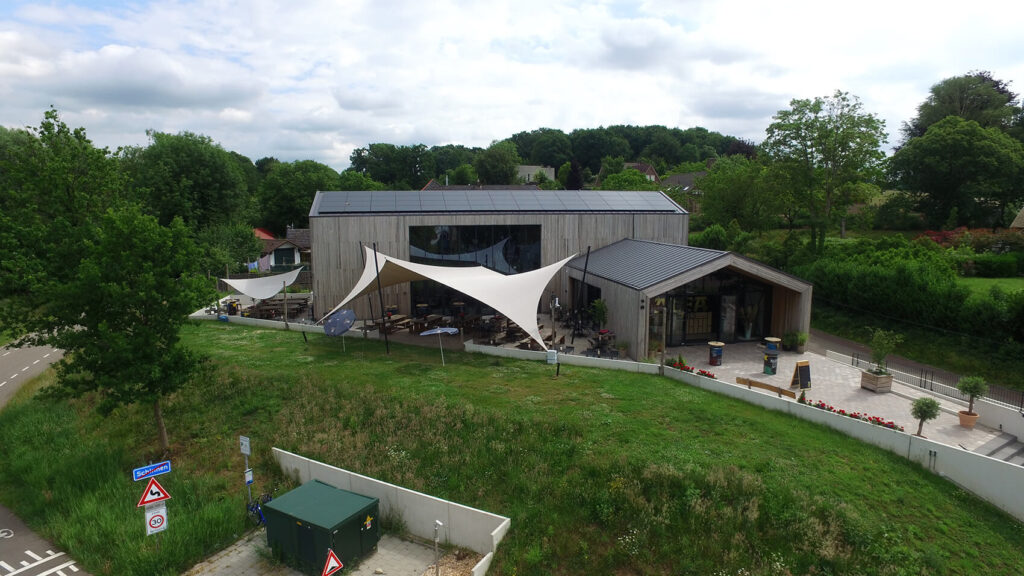 A modern wooden building with solar panels and an outdoor seating area under white shade sails beside a green field.