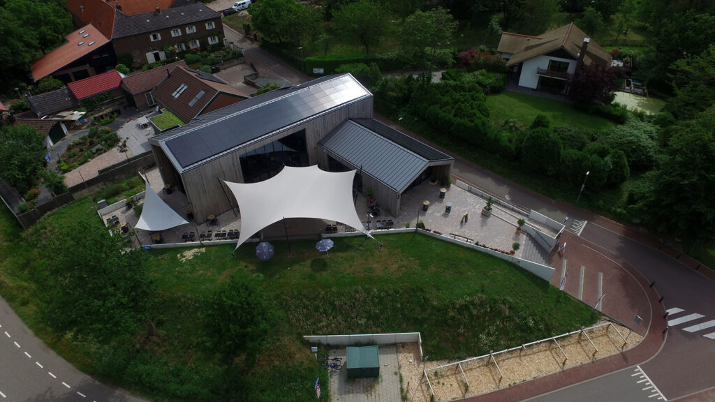 Aerial view of a modern wooden building with a large white sail-shaped canopy and outdoor seating on a paved patio adjacent to a green lawn and road.