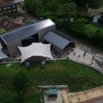 Aerial view of a modern wooden building with a large white sail-shaped canopy and outdoor seating on a paved patio adjacent to a green lawn and road.