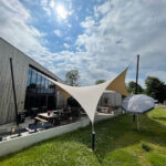Outdoor cafe patio with wooden tables beneath a white tensile canopy next to a metal-clad building, under a bright blue sky with sun.