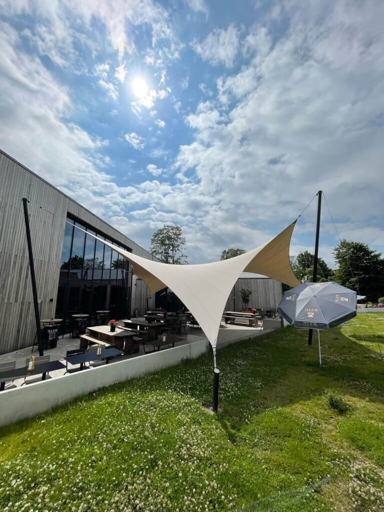 Outdoor cafe patio with wooden tables beneath a white tensile canopy next to a metal-clad building, under a bright blue sky with sun.