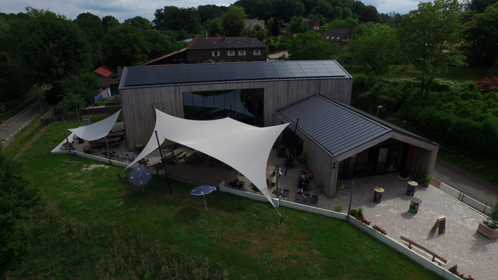 Aerial view of a modern wooden building with white fabric shade sails over an outdoor patio and seating area surrounded by greenery and a paved courtyard.