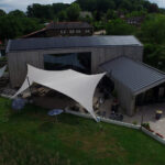 Aerial view of a modern wooden building with white fabric shade sails over an outdoor patio and seating area surrounded by greenery and a paved courtyard.