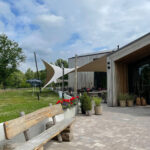 Outdoor cafe or restaurant terrace with fabric shade sails, wooden building, and potted plants along a tiled patio.