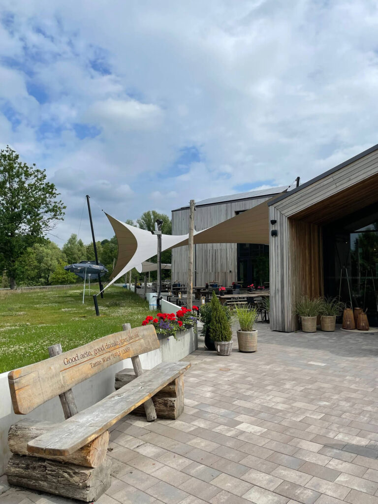 Outdoor cafe or restaurant terrace with fabric shade sails, wooden building, and potted plants along a tiled patio.