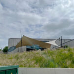 Contemporary building with tan and white fabric shade sails over an outdoor seating area on a grassy slope under a cloudy sky.
