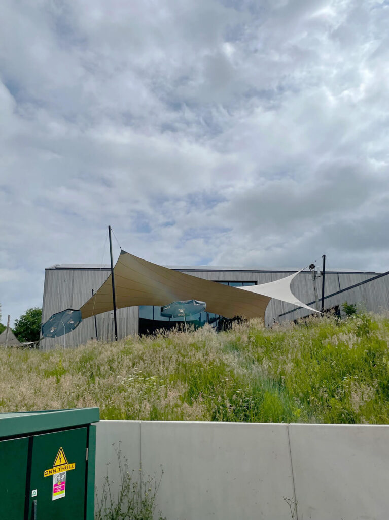 Contemporary building with tan and white fabric shade sails over an outdoor seating area on a grassy slope under a cloudy sky.