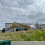 Modern gray building with tan and white fabric shade sails and blue patio umbrellas over a seating area, with tall grasses in the foreground.