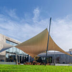 Modern building with wooden cladding and large glass doors, shaded by a beige fabric sail over an outdoor seating area.