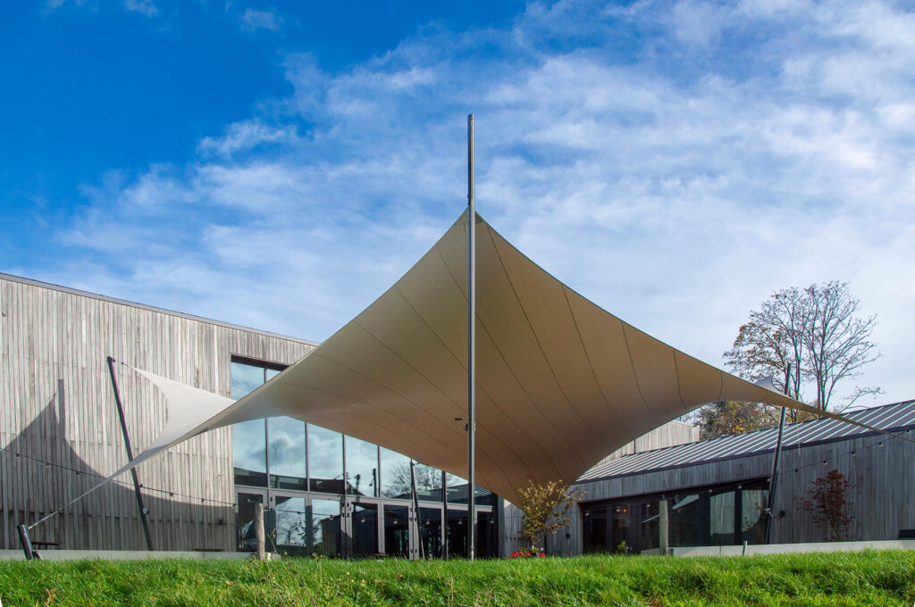 Modern building with wood cladding and a large beige fabric shade sail held by a central pole against a blue sky.