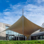Modern building with wood cladding and a large beige fabric shade sail held by a central pole against a blue sky.