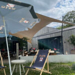 Outdoor cafe seating with large fabric shade sails and a blue ALFA-branded deck chair in foreground; a person sits at a table in the background.