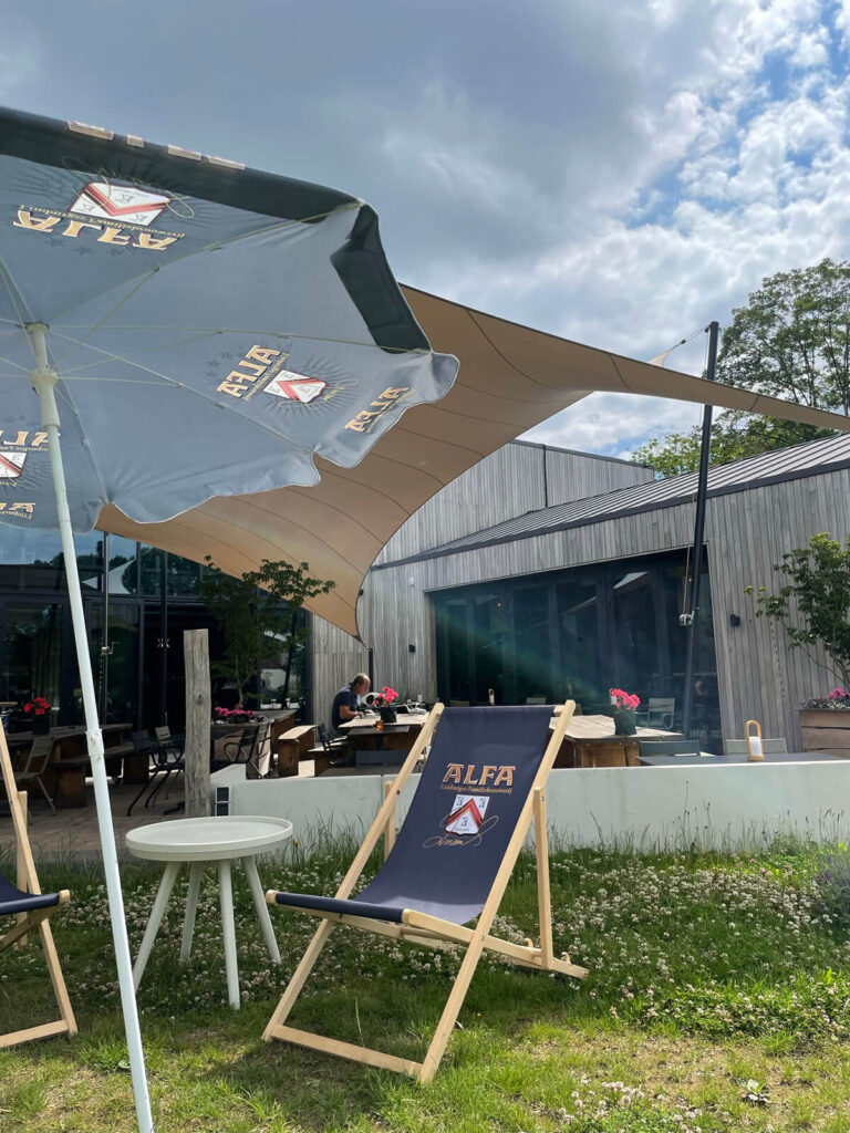 Outdoor cafe seating with large fabric shade sails and a blue ALFA-branded deck chair in foreground; a person sits at a table in the background.