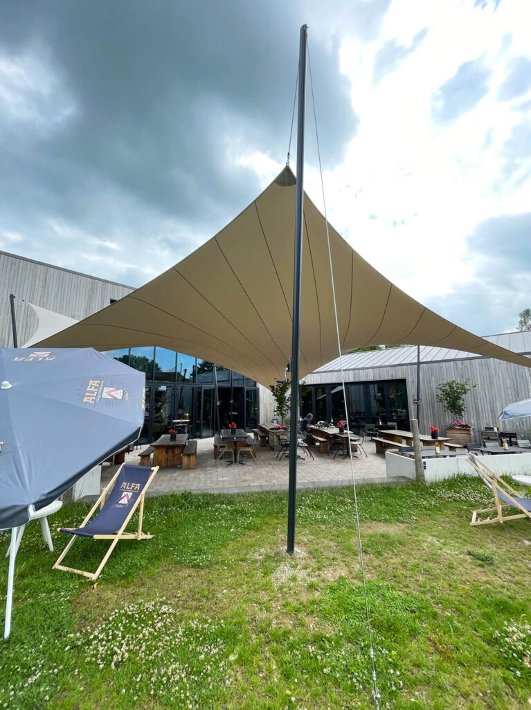 Outdoor cafe patio under a large beige shade sail, with wooden tables and benches and a modern building in the background.